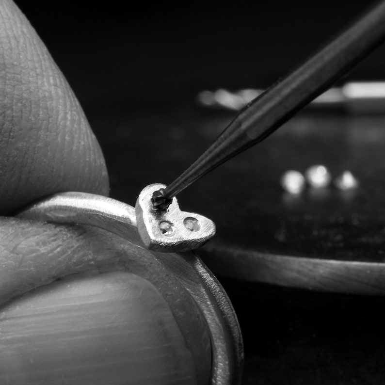 May include: A silver ring featuring a heart-shaped setting is being worked on with a precision tool. The heart element contains two small, circular indentations, and a third indentation is being carefully created by the tip of the metal tool.