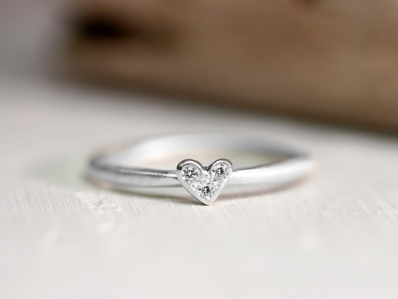 May include: A silver-toned ring featuring a small heart-shaped centerpiece. The heart is embellished with three tiny, clear, round-cut stones arranged in a triangular pattern. The band has a smooth, matte finish and a rounded profile.