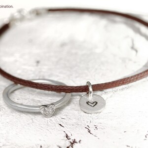 May include: A brown cord bracelet featuring a silver ring with a small heart-shaped accent and a round silver charm engraved with a heart outline. The bracelet is displayed with the text "Beispiel Kombination" in the upper left corner.
