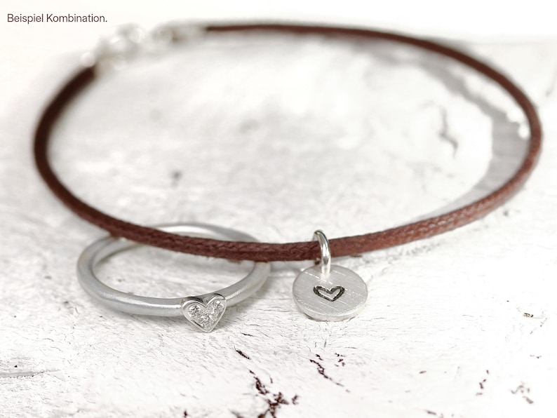 May include: A brown cord bracelet featuring a silver ring with a small heart-shaped accent and a round silver charm engraved with a heart outline. The bracelet is displayed with the text "Beispiel Kombination" in the upper left corner.