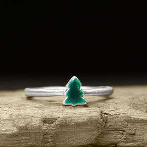 Könnte beinhalten: Ein silberner Ring mit einem kleinen, grünen Emaille-Weihnachtsbaum-Design. Der Ring ist vor einem neutralen Hintergrund platziert, der die Details hervorhebt. Der Ring ist ein Schmuckstück.
