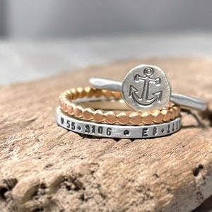 May include: Three silver rings stacked on a wooden surface. The top ring features an anchor design. The middle ring is a beaded band. The bottom ring has the coordinates "N 55.3106 E 8.2410" engraved on it.
