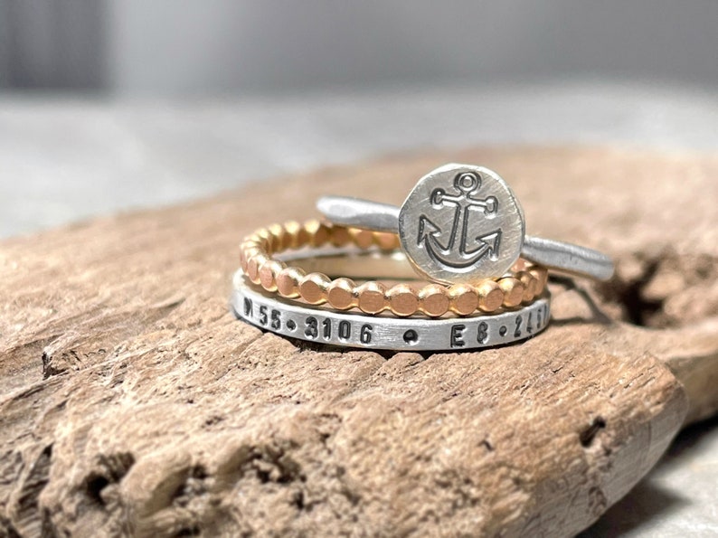 May include: Three silver rings stacked on a wooden surface. The top ring features an anchor design. The middle ring is a beaded band. The bottom ring has the coordinates "N 55.3106 E 8.2410" engraved on it.