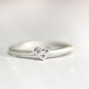 May include: A silver-toned ring featuring a small heart-shaped centerpiece. The heart is embellished with three tiny, clear, sparkling stones arranged in a triangular pattern. The band of the ring is smooth, rounded, and has a polished metallic finish.