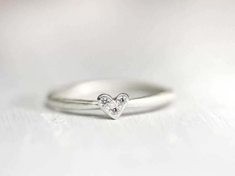 May include: A silver-toned ring featuring a small heart-shaped centerpiece. The heart is embellished with three tiny, clear, sparkling stones arranged in a triangular pattern. The band of the ring is smooth, rounded, and has a polished metallic finish.