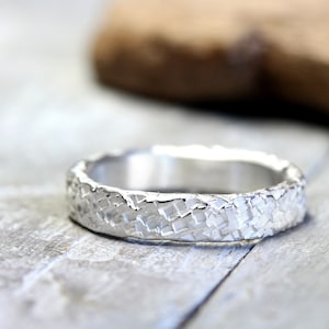 Könnte beinhalten: Ein silberner Ring mit strukturierter Oberfläche. Der Ring hat ein Muster aus kleinen, erhabenen Quadraten, was ihm ein raues, handgefertigtes Aussehen verleiht. Der Ring wird auf einer hellgrauen Oberfläche gezeigt. Das Design des Rings ist einzigartig.