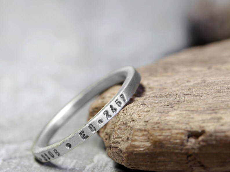 May include: A silver ring with a series of numbers engraved around the band. The ring is resting on a piece of weathered wood.