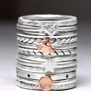 May include: A stack of silver rings featuring various textures and charms, including a heart with small clear stones, a rose gold paper boat, a silver star, and a rose gold circular disc. The bands vary between smooth, hammered, and twisted designs.
