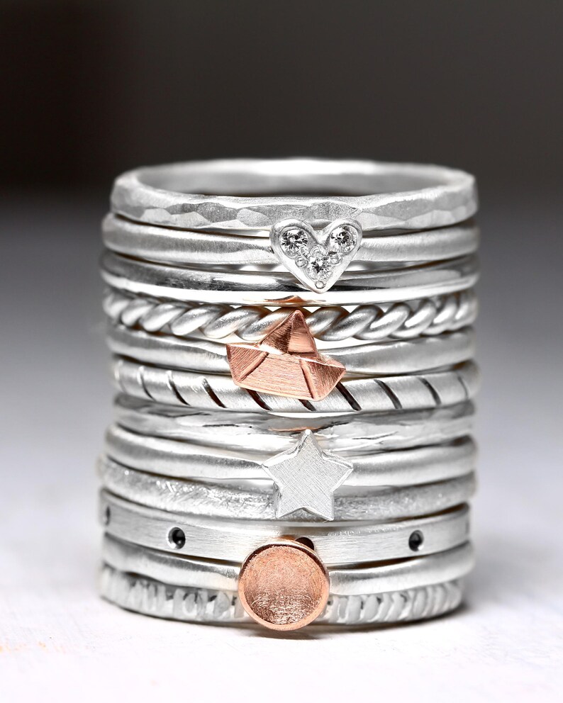 May include: A stack of silver rings featuring various textures and charms, including a heart with small clear stones, a rose gold paper boat, a silver star, and a rose gold circular disc. The bands vary between smooth, hammered, and twisted designs.