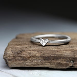 May include: A silver-toned ring featuring a small heart-shaped centerpiece. The heart is embellished with three tiny, clear, sparkling stones arranged in a triangular pattern. The band has a smooth, matte finish and a rounded, consistent profile.