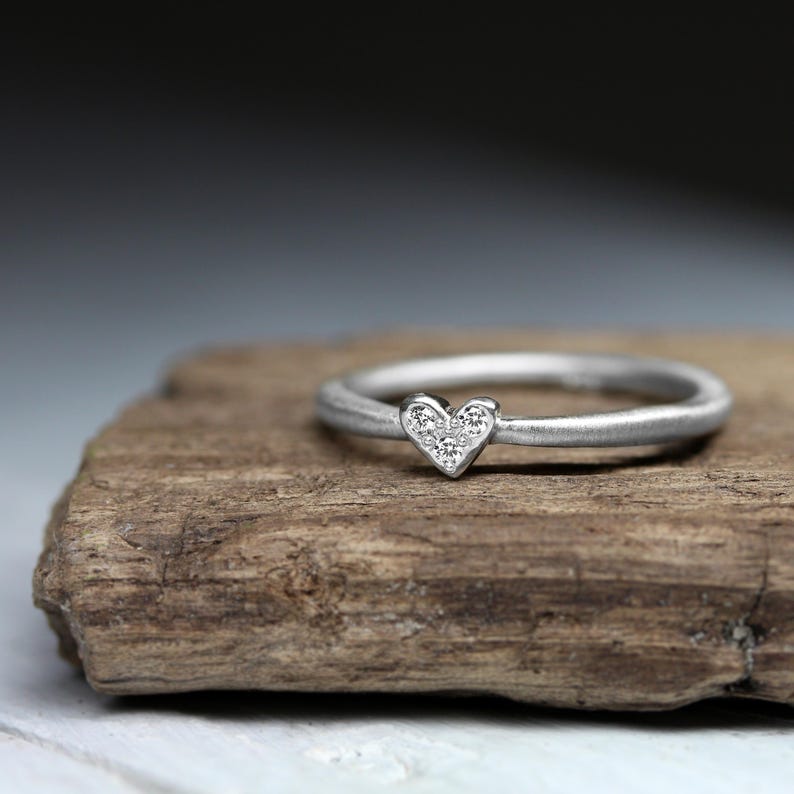 May include: A silver-toned ring featuring a small heart-shaped centerpiece. The heart is embellished with three tiny, clear, sparkling stones arranged in a triangular pattern. The band has a smooth, matte finish and a rounded, consistent profile.