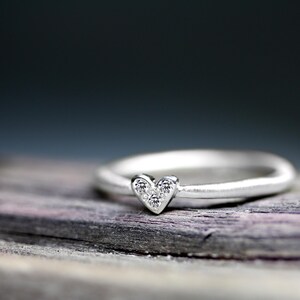 May include: A silver ring featuring a small heart-shaped setting at the center. The heart-shaped element is inlaid with three tiny, clear, sparkling stones arranged in a triangular pattern, creating a delicate and refined appearance on the band.