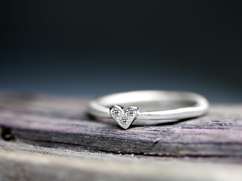 May include: A silver ring featuring a small heart-shaped setting at the center. The heart-shaped element is inlaid with three tiny, clear, sparkling stones arranged in a triangular pattern, creating a delicate and refined appearance on the band.