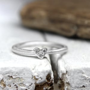 May include: A silver-toned ring featuring a small heart-shaped centerpiece. The heart is embellished with three tiny, clear, sparkling stones arranged in a triangular pattern. The band of the ring has a smooth, matte, metallic finish.