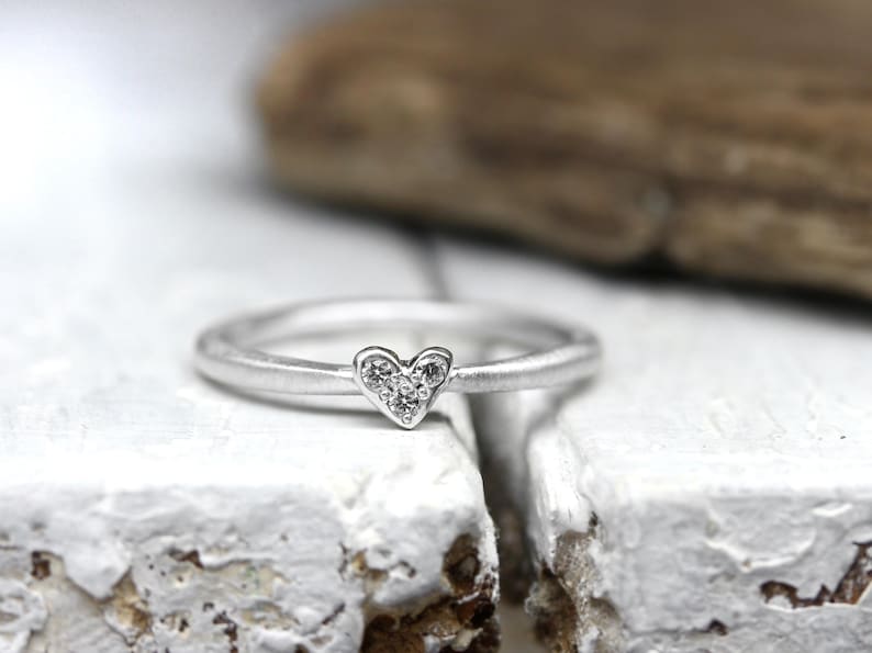 May include: A silver-toned ring featuring a small heart-shaped centerpiece. The heart is embellished with three tiny, clear, sparkling stones arranged in a triangular pattern. The band of the ring has a smooth, matte, metallic finish.