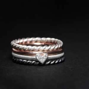 May include: A stack of four rings featuring a silver twisted band, a smooth rose gold band, a matte silver band with a small heart-shaped charm set with three tiny clear stones, and a dark grey twisted band, all resting against a black background.