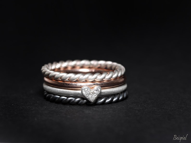 May include: A stack of four rings featuring a silver twisted band, a smooth rose gold band, a matte silver band with a small heart-shaped charm set with three tiny clear stones, and a dark grey twisted band, all resting against a black background.