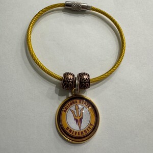 May include: A gold-colored keychain with a circular pendant featuring the Arizona State University logo. The pendant has a gold border, a white background, and a maroon pitchfork emblem. The keychain has a gold wire loop and decorative beads.