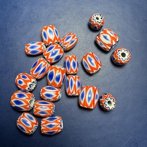May include: A collection of 17 colorful glass beads with a white, orange, and blue geometric pattern. The beads are oval shaped and have a hole in the center.