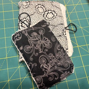 May include: Two fabric items with floral patterns. One is black and gray with a swirling design, the other is white and black with a floral pattern. Both have a white, fluffy border and a black elastic band.