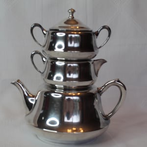 May include: A three-piece silver tea set. The set includes a teapot, a creamer, and a sugar bowl with a lid. The pieces are stacked on top of each other. The set is made of a shiny, reflective metal and sits on a white surface.