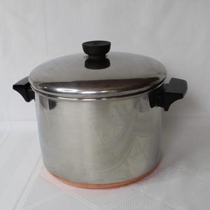May include: A stainless steel stockpot with a matching lid, featuring black plastic handles on the sides and a black knob on the lid. The base of the pot is constructed with a visible copper-colored band, providing a distinct contrast to the silver-toned metal body of the cooking vessel.