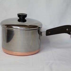 May include: A stainless steel saucepan with a copper-colored base, a matching stainless steel lid with a black knob, and a black handle featuring a small metal hanging loop at the end. The pot has a cylindrical body and a polished metallic finish.
