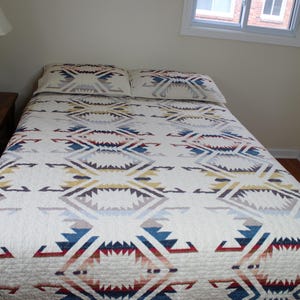 May include: A bed with a white quilt and two matching pillowcases. The quilt features a geometric pattern in shades of blue, red, yellow, and gray. The bed is in a room with a window and a neutral-colored wall.
