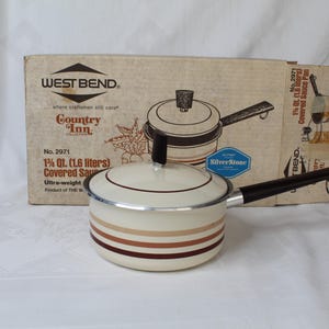 May include: Cream-colored West Bend Country Inn covered saucepan with brown stripes and a black handle. The original box is behind it, featuring an illustration of the pan and the text "SilverStone". The pan is 1.6 liters.