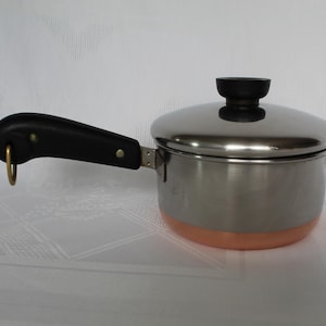 May include: A small stainless steel saucepan with a copper-colored base and a black handle. The saucepan has a lid with a black knob and a metal ring on the handle for hanging. The pot is designed for cooking.