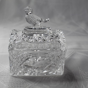 May include: A clear, square glass trinket box with a bird-shaped lid. The box features a detailed, geometric pattern and an etched bird design on the front. The lid is topped with a detailed bird figurine.
