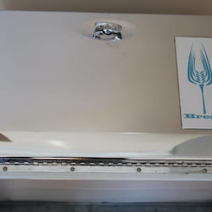 May include: A shiny, chrome bread box with a hinged lid and a decorative handle. A rectangular label on the front features a blue wheat stalk design and the word "Bread". The box is designed to store bread.