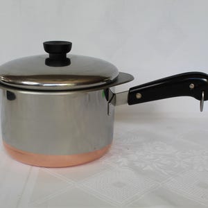 May include: Stainless steel saucepan with a copper-colored base and a black handle. The pot has a matching lid with a black knob. The handle has a metal ring at the end. The pot is on a white surface.