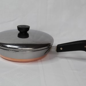 May include: A stainless steel frying pan with a copper-colored base and a black handle. The pan has a lid with a black knob. The handle has a hole for hanging. Kitchen cookware.