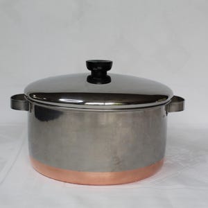 May include: A stainless steel cooking pot with a copper-colored base and a shiny lid. The pot has two handles and a black knob on top of the lid. The pot is designed for cooking and food preparation.