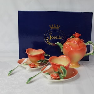 May include: A Sorelle Fine Porcelain tea set featuring a teapot and two teacups with saucers. The set is orange and yellow with green accents. The teacups are heart-shaped. The set is presented in a navy blue box.