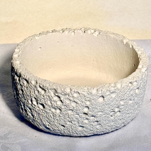 May include: A small, round, off-white ceramic bowl with a textured, porous exterior. The bowl's rim has an uneven, slightly jagged edge. The interior is smooth and light-colored. The bowl is likely used for decorative purposes or as a small planter.