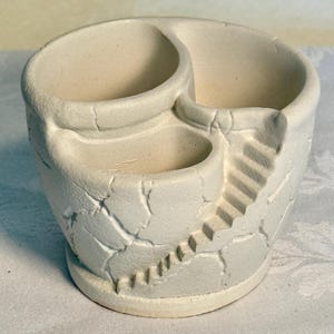 May include: Off-white ceramic planter with three compartments and a textured, cracked surface. The planter features a small staircase design on the side, adding a unique architectural element. The compartments are of varying sizes, suitable for small plants or decorative items.
