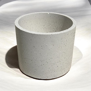 May include: A cylindrical, light gray concrete planter with a speckled texture. The planter has a smooth, rounded rim and a simple, modern design. The planter is suitable for small plants and succulents.