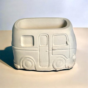 May include: A small, off-white planter shaped like a vintage camper van. The planter features detailed molding of windows, doors, and wheels. The rectangular opening at the top is designed for planting small succulents or flowers.