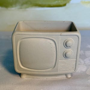 May include: A small, off-white ceramic planter shaped like a vintage television set. The planter has a rectangular opening, a rounded screen, two circular knobs, and a speaker grill design on the side. It sits on two small feet.