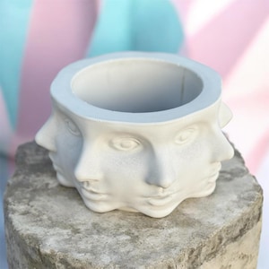 May include: A white, multi-faced planter with a circular opening. The planter is made of a smooth, matte material and features three visible faces. It sits on a rough, gray stone surface, with a pastel pink and blue background.