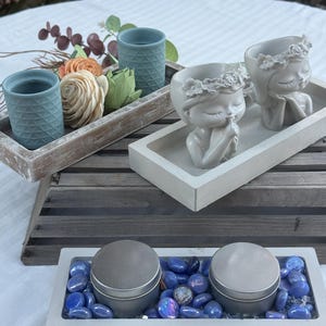 May include: A collection of decorative items, including small blue textured ceramic cups, floral arrangements, and two gray containers with lids, displayed on wooden and concrete trays. The scene is set on a white tablecloth.