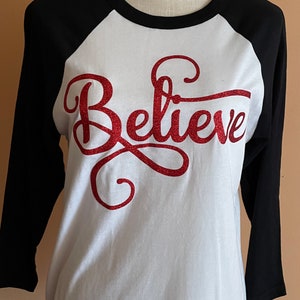 May include: White and black baseball tee shirt with red glitter lettering that says "Believe".