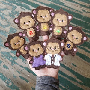 May include: Set of seven brown felt finger puppets shaped like monkeys. Each monkey has a different embroidered design, including numbers 1 through 5, a doctor's stethoscope, and a green leaf.