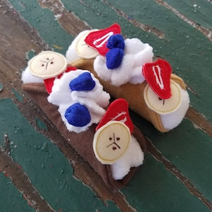 May include: Two handmade felt cannoli, one with chocolate and one with tan filling. Both are topped with white felt frosting, red felt strawberries, and blue felt blueberries.