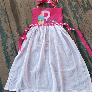May include: Pink and white gingham fabric apron with a gumball machine and the letter "P" embroidered on the front. The apron has white polka dot trim and ties. The text "Penny" is embroidered below the gumball machine.