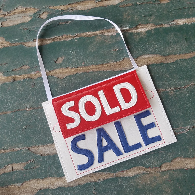 May include: A white sign with red and blue fabric lettering that reads "SOLD SALE". The sign has a white loop at the top for hanging.