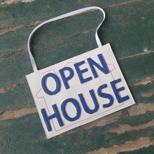 May include: White fabric sign with a blue stitched outline of a house and the words "OPEN HOUSE" in blue stitched letters.