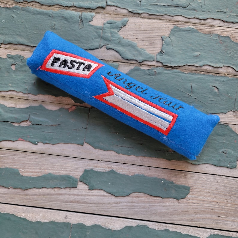 May include: Blue felt tube with red and white embroidered details. The tube has the word "PASTA" and the phrase "Angel Hair" embroidered on it.